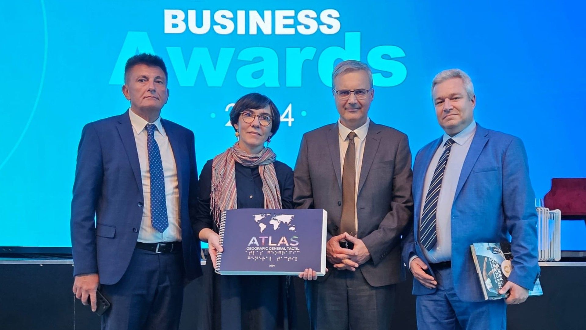 Fotografie de la ceremonia de decernare a premiilor Business Awards. Un grup de patru persoane stă în fața unei săli festive și țin în mijlocul lor Atlasul Tactil. Toți privesc spre fotograf și zâmbesc. De la stânga la dreapta, aceștia sunt: Dorin Miron, președintele Clubului Francofon de Afaceri din Timișoara, Alina Satmari, coordonatorul proiectului Atlas Tactil, Excelența sa Nicolas Warnery, Ambasadorul Franței în România, și Mădălin Bunoiu, prorector al Universității de Vest din Timișoara. Fotografie este realizată după momentul în care Atlasul Tactil a primit premiul pentru inovare pe anul 2024.